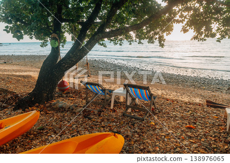 ellow kayaks and relaxing beach chairs under a large tree on a tranquil tropical pebble beach at sunset. 138976065
