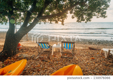 ellow kayaks and relaxing beach chairs under a large tree on a tranquil tropical pebble beach at sunset. ellow kayaks and relaxing beach chairs under a large tree on a tranquil tropical pebble beach at sunset. 138976066