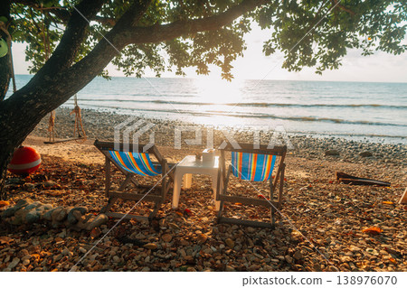 Relaxing Beach Chairs Under Tree Shade at Sunrise on a Pebbly Shore Relaxing Beach Chairs Under Tree Shade at Sunrise on a Pebbly Shore 138976070