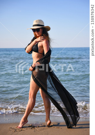 Woman walking on beach in black bikini with sheer cover up and sun hat Woman walking on beach in black bikini with sheer cover up and sun hat 138976342