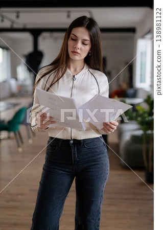 Young professional woman carefully examining business documents while standing in a modern office, focusing on financial reports, contracts, or project plans Young professional woman carefully examining business documents while standing in a modern office, focusing on financial reports, contracts, or project plans 138981122