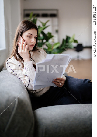 Young woman working from home on a sofa, holding documents and speaking intently on the phone while reviewing paperwork and planning business details during a remote work call Young woman working from home on a sofa, holding documents and speaking intently on the phone while reviewing paperwork and planning business details during a remote work call 138981124