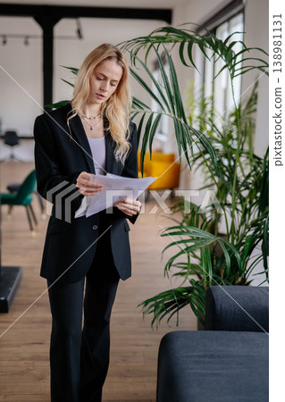Businesswoman reviewing important business documents and contracts, standing focused amidst a contemporary office setting, representing professional career management and corporate success 138981131