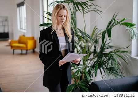 Young blonde woman in a black blazer stands in a modern office, intently reviewing paperwork and contracts, focused on corporate documents and strategic planning for her role Young blonde woman in a black blazer stands in a modern office, intently reviewing paperwork and contracts, focused on corporate documents and strategic planning for her role 138981132