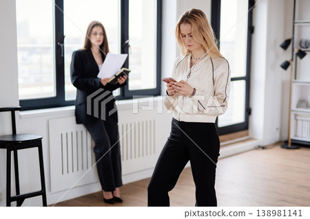 Two businesswomen standing in a modern office space, one woman focusing on her smartphone while the other reads documents by a large window, both engaged in their work Two businesswomen standing in a modern office space, one woman focusing on her smartphone while the other reads documents by a large window, both engaged in their work 138981141
