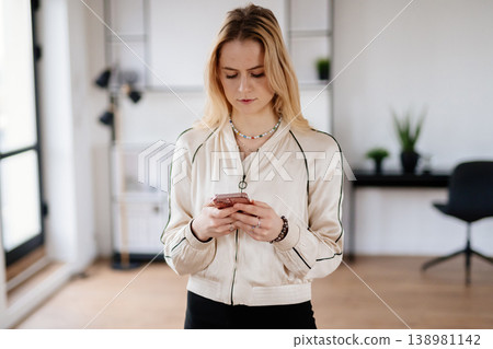Young blonde woman standing indoors, focused on interacting with a smartphone, representing communication, social networking, and digital connection in a modern setting 138981142