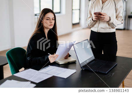 Young businesswoman at a desk carefully reviewing documents while a colleague stands nearby checking a smartphone, conveying teamwork, focus and modern office productivity 138981143