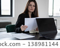 Young professional businesswoman at a modern desk, focused on paperwork and a laptop, analyzing reports with pen in hand in a bright office, concentrating on career tasks 138981144