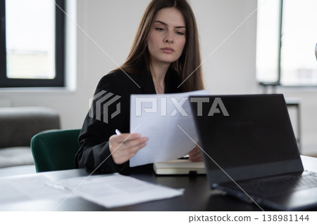 Young professional businesswoman at a modern desk, focused on paperwork and a laptop, analyzing reports with pen in hand in a bright office, concentrating on career tasks Young professional businesswoman at a modern desk, focused on paperwork and a laptop, analyzing reports with pen in hand in a bright office, concentrating on career tasks 138981144