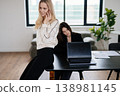 Two women professionals concentrating on work, one making a phone call while the other works on a laptop at a table, embodying teamwork and communication in a contemporary business setting 138981145