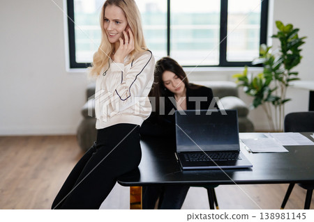Two women professionals concentrating on work, one making a phone call while the other works on a laptop at a table, embodying teamwork and communication in a contemporary business setting 138981145
