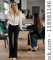 Young businesswoman on a phone call stands at a conference table while colleague seated beside her takes notes on a laptop and notebook during a focused office meeting 138981146