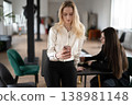 Blonde businesswoman checking her mobile phone, using social media or communicating with clients while standing beside a desk, collaborating with a colleague in a bright office space 138981148