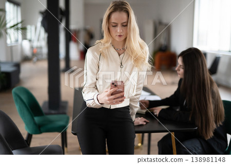 Blonde businesswoman checking her mobile phone, using social media or communicating with clients while standing beside a desk, collaborating with a colleague in a bright office space Blonde businesswoman checking her mobile phone, using social media or communicating with clients while standing beside a desk, collaborating with a colleague in a bright office space 138981148