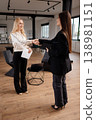 Two smiling businesswomen shaking hands in a modern office space, finalizing an agreement and demonstrating successful partnership, collaboration, and professional greeting 138981151