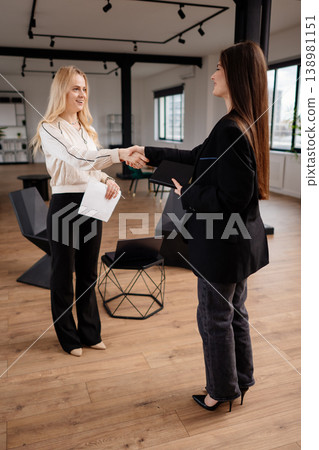Two smiling businesswomen shaking hands in a modern office space, finalizing an agreement and demonstrating successful partnership, collaboration, and professional greeting Two smiling businesswomen shaking hands in a modern office space, finalizing an agreement and demonstrating successful partnership, collaboration, and professional greeting 138981151