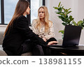 Two young women collaborating on a project, one typing on a laptop and another holding a notebook while engaged in a focused business discussion in a modern office lounge setting 138981155