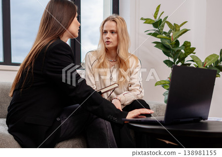 Two young women collaborating on a project, one typing on a laptop and another holding a notebook while engaged in a focused business discussion in a modern office lounge setting Two young women collaborating on a project, one typing on a laptop and another holding a notebook while engaged in a focused business discussion in a modern office lounge setting 138981155