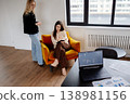 Two businesswomen collaborate in a modern office, one standing and one seated in an armchair reviewing notes and a laptop with charts for project planning and strategy discussion 138981156
