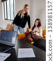 Two young women colleagues are working together, discussing new ideas and taking notes in a relaxed open plan office environment, fostering teamwork and professional communication 138981157