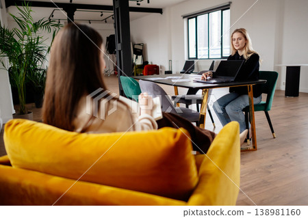 Two women engaging in a professional business meeting, one taking notes while the other uses a laptop, fostering discussion and collaboration in a contemporary coworking space Two women engaging in a professional business meeting, one taking notes while the other uses a laptop, fostering discussion and collaboration in a contemporary coworking space 138981160