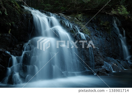 A waterfall flowing down a black rock face in multiple streams; the trails of water captured by long exposure, and the silence of the forest. 138981714
