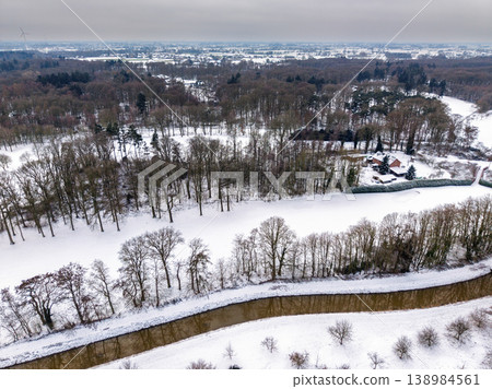 Aerial winter landscape featuring a winding river flowing through snow-covered fields and forests. A peaceful countryside view with bare trees, a lonely house, and distant windmills under a grey sky. 138984561