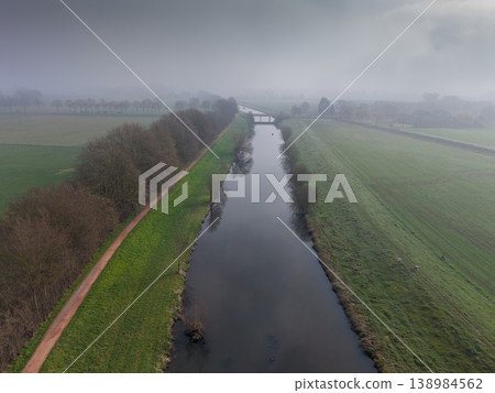 Aerial view of a peaceful river canal on a foggy morning. A dirt path runs alongside green fields and bare trees, disappearing into thick mist. Moody and tranquil rural landscape in Europe. 138984562