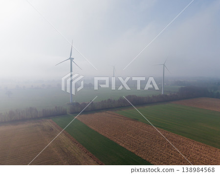 Aerial view of wind turbines rising through thick morning fog over agricultural fields. A sustainable energy concept featuring a misty, atmospheric rural landscape at dawn in Europe. Aerial view of wind turbines rising through thick morning fog over agricultural fields. A sustainable energy concept featuring a misty, atmospheric rural landscape at dawn in Europe. 138984568