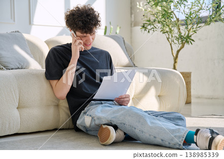 Young handsome guy, 20s university student talking on cell phone with paper documents Young handsome guy, 20s university student talking on cell phone with paper documents 138985339