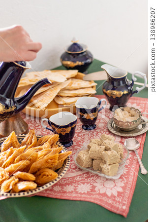Hand pours coffee from a cobalt blue vintage decanter pot in cup. Sweets food on table honey baklava, halva on a patterned red tablecloth Hand pours coffee from a cobalt blue vintage decanter pot in cup. Sweets food on table honey baklava, halva on a patterned red tablecloth 138986207