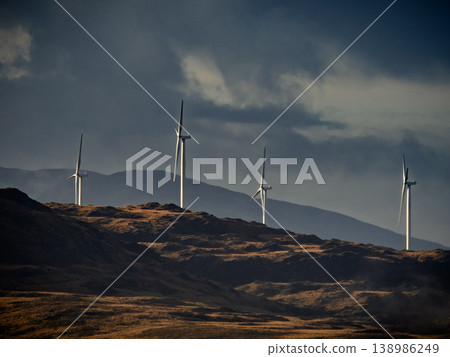 tall wind turbines stand on a dark, rugged hillside generating renewable energy under a dramatic, cloudy sky. 138986249
