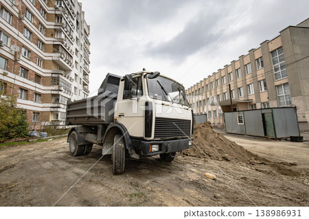 A truck is parked on a dirt area next to a construction site. Nearby buildings show signs of urban development. The sky is cloudy above the scene A truck is parked on a dirt area next to a construction site. Nearby buildings show signs of urban development. The sky is cloudy above the scene 138986931