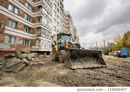A construction vehicle operates near an apartment building. Workers are involved in site preparation. Clouds cover the sky on this urban day 138986947