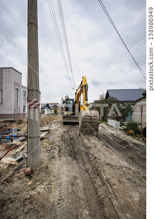 Excavation takes place on a residential street where a large machine works on the ground near utility poles and houses Excavation takes place on a residential street where a large machine works on the ground near utility poles and houses 138986949
