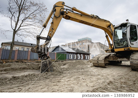 Construction is happening as an excavator digs into the ground near several buildings under a gray sky in an urban area Construction is happening as an excavator digs into the ground near several buildings under a gray sky in an urban area 138986952