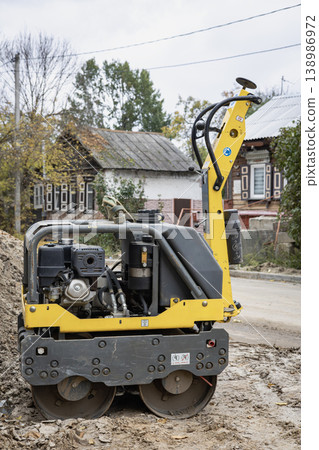 A yellow construction machine sits at a road work site in a small town. The machine is designed for rolling and compacting asphalt or soil A yellow construction machine sits at a road work site in a small town. The machine is designed for rolling and compacting asphalt or soil 138986972