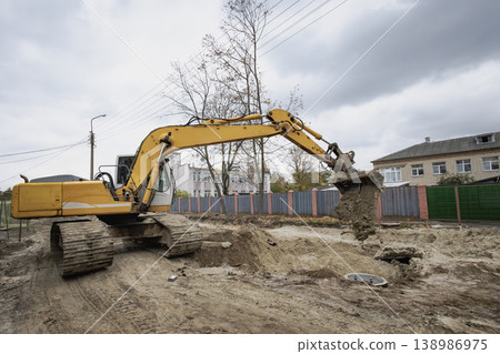 A yellow excavator works on the soil at a construction site in a town. It is cloudy and there are buildings in the background A yellow excavator works on the soil at a construction site in a town. It is cloudy and there are buildings in the background 138986975