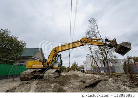 A yellow excavator digs in the ground at a construction site in a residential neighborhood on a cloudy afternoon. Trees and buildings are nearby 138986983