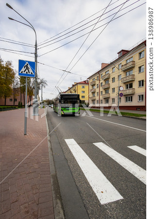 A bus moves along the road next to a crosswalk and apartments on a cloudy autumn day. People walk on the sidewalk nearby while trees line the street A bus moves along the road next to a crosswalk and apartments on a cloudy autumn day. People walk on the sidewalk nearby while trees line the street 138986987