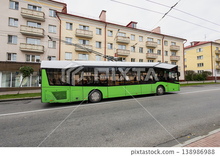 A green city bus drives down the street beside several apartment buildings. The sky is overcast, and the street is quiet A green city bus drives down the street beside several apartment buildings. The sky is overcast, and the street is quiet 138986988