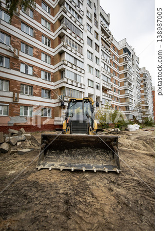 A backhoe loader is on a construction site in front of a tall apartment building. Workers are preparing the ground for a new project in the area A backhoe loader is on a construction site in front of a tall apartment building. Workers are preparing the ground for a new project in the area 138987005