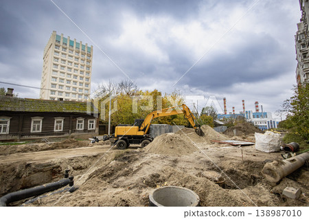 Heavy machinery is digging in the ground at a construction site near a tall building and old houses on a cloudy day in the city 138987010