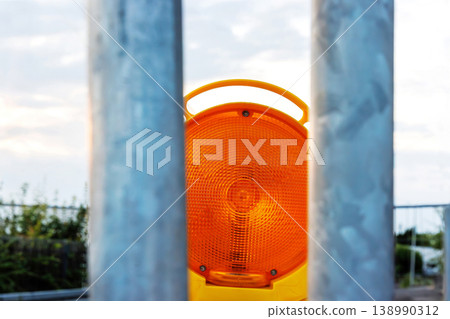 Orange warning beacon framed by metal fence bars at roadwork site against cloudy sky background with copy space Orange warning beacon framed by metal fence bars at roadwork site against cloudy sky background with copy space 138990312