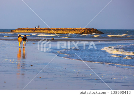 Couple walking hand in hand on a wet sandy beach during golden hour sunset Couple walking hand in hand on a wet sandy beach during golden hour sunset 138990413