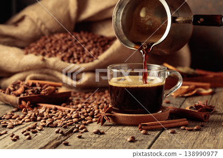 Black coffee is poured from a copper coffee pot into a glass cup. 138990977