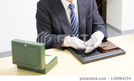 A staff member at a buyback shop places a watch and a branded wallet on a tray and appraises them. A staff member at a buyback shop places a watch and a branded wallet on a tray and appraises them. 138994254