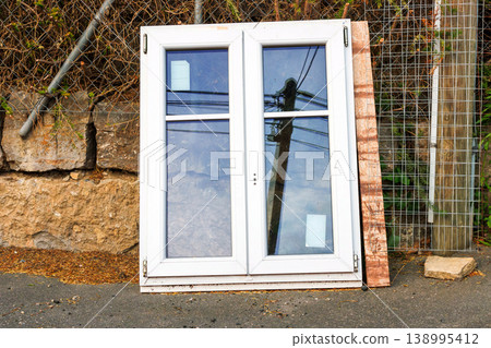 White plastic window frame with glass panes leans against a stone wall beside a road. Discarded building material and renovation debris. White plastic window frame with glass panes leans against a stone wall beside a road. Discarded building material and renovation debris. 138995412