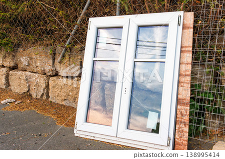 White plastic window frame with glass panes leans against a stone wall beside a road. Discarded building material and renovation debris. White plastic window frame with glass panes leans against a stone wall beside a road. Discarded building material and renovation debris. 138995414