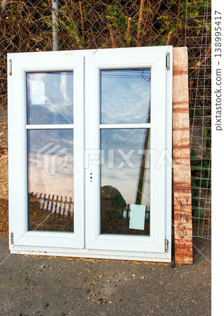 White plastic window frame with glass panes leans against a stone wall beside a road. Discarded building material and renovation debris. White plastic window frame with glass panes leans against a stone wall beside a road. Discarded building material and renovation debris. 138995417